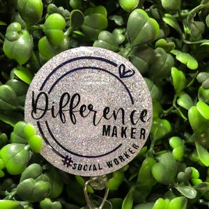 May include: A round, glittery badge reel with the words "Difference Maker" in black script. The text is encircled by a black line, with a small heart symbol. The hashtag #SOCIAL WORKER is also included. The background is a bed of green leaves.