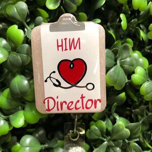 May include: A badge reel with a clipboard design. The clipboard has the words "HIM Director" in red, a red heart, and a stethoscope. The badge reel is attached to a clear plastic holder with a decorative, glittery button.