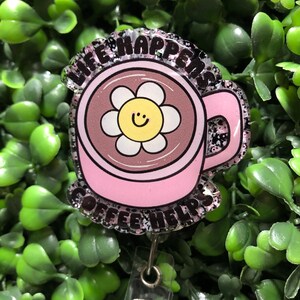 May include: A pink coffee cup-shaped badge reel with the text "LIFE HAPPENS COFFEE HELPS". The cup features a daisy with a smiling face. The badge reel has a clear plastic retractable cord and a silver-colored clip.
