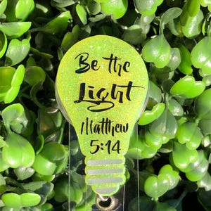 May include: A green glitter lightbulb shaped badge holder with the text "Be the light Matthew 5:14" and a clear retractable badge holder with a sparkly iridescent charm.