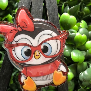 May include: A badge reel featuring a cartoon penguin wearing red glasses and a red bow. The penguin is dressed in a red sweater and has orange feet. The badge reel has a glittery border and the text "New Designs Studio".
