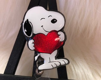 May include: A badge reel featuring a white cartoon dog with a black ear, winking and holding a large, glittery red heart. The character is attached to a clear plastic strap with a small, round, iridescent, glittery accent button at the base.