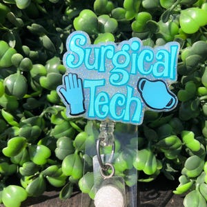 May include: A light blue badge reel with the words "Surgical Tech" in a whimsical font. The badge features a blue glove and a surgical mask. The background is a lush green plant.