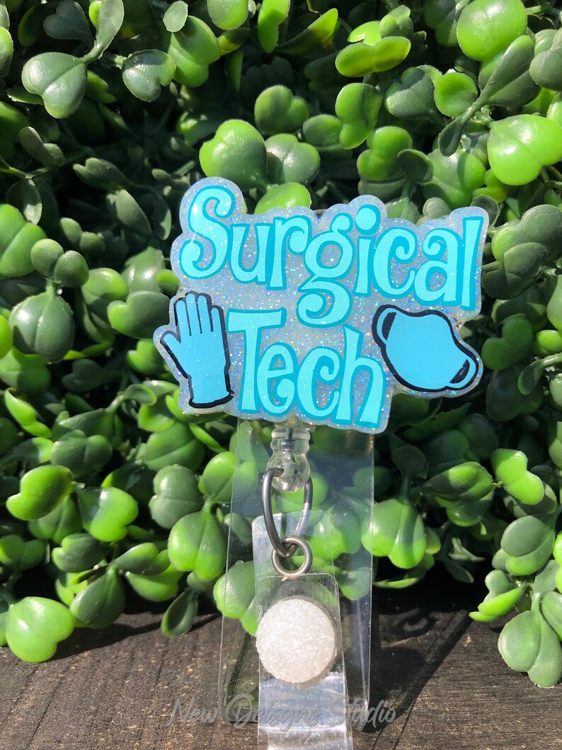 May include: A light blue badge reel with the words "Surgical Tech" in a whimsical font. The badge features a blue glove and a surgical mask. The background is a lush green plant.