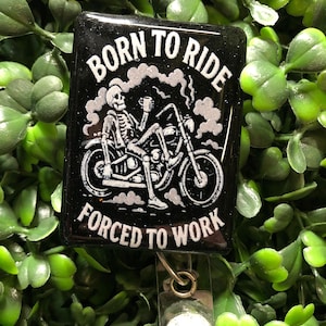 May include: Black rectangular badge reel with the text "BORN TO RIDE FORCED TO WORK" and a skeleton riding a motorcycle. The design is in white against a black background. The reel has a clear plastic strap and a metal clip.