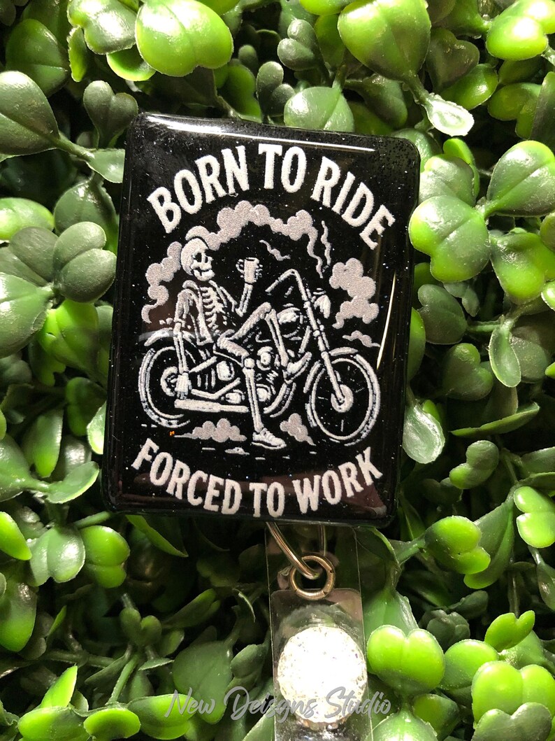 May include: Black rectangular badge reel with the text "BORN TO RIDE FORCED TO WORK" and a skeleton riding a motorcycle. The design is in white against a black background. The reel has a clear plastic strap and a metal clip.
