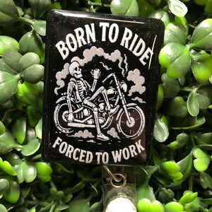 May include: Black badge with the text "BORN TO RIDE FORCED TO WORK" and a skeleton riding a motorcycle. The badge has a clear plastic holder and a metal clip. The background is green.