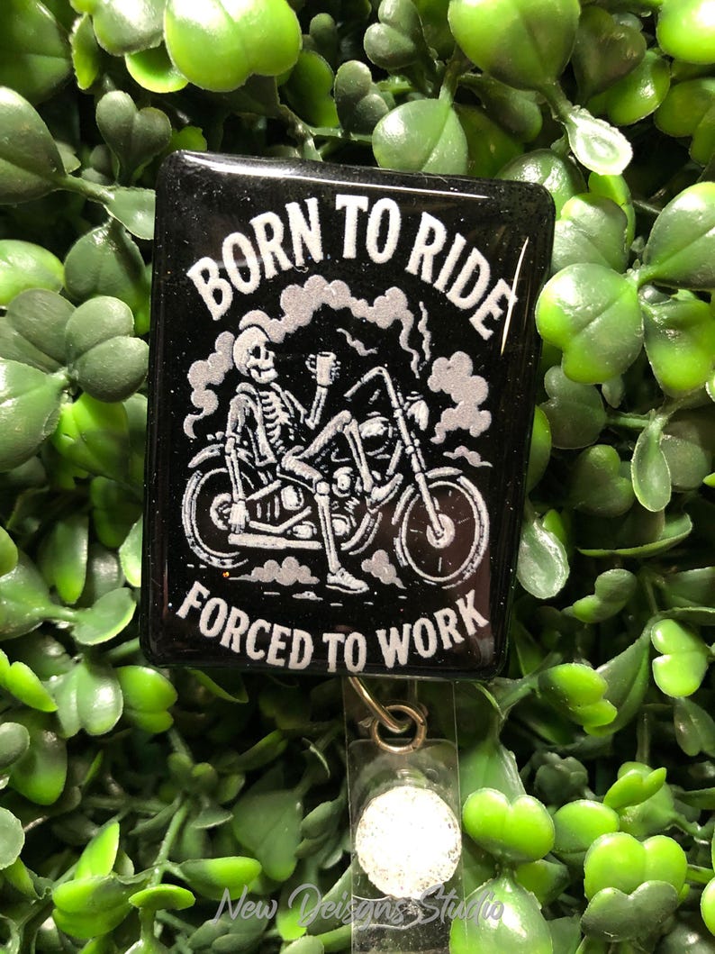 May include: Black badge with the text "BORN TO RIDE FORCED TO WORK" and a skeleton riding a motorcycle. The badge has a clear plastic holder and a metal clip. The background is green.