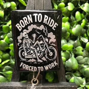 May include: Black badge with the text "BORN TO RIDE FORCED TO WORK" and a skeleton riding a motorcycle. The design is in white against a black background. The badge is rectangular and has a clear plastic holder.
