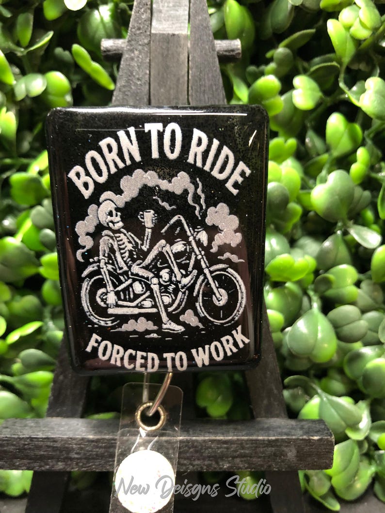 May include: Black badge with the text "BORN TO RIDE FORCED TO WORK" and a skeleton riding a motorcycle. The design is in white against a black background. The badge is rectangular and has a clear plastic holder.