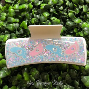 Pu&ograve; includere: Un fermaglio per capelli rettangolare bianco con un centro glitterato e iridescente. La clip presenta disegni di impronte di piedi di bambino blu e rosa. Il testo "New Designs Studio" &egrave; visibile in basso.