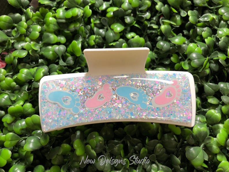 Pu&ograve; includere: Un fermaglio per capelli rettangolare bianco con un centro glitterato e iridescente. La clip presenta disegni di impronte di piedi di bambino blu e rosa. Il testo "New Designs Studio" &egrave; visibile in basso.