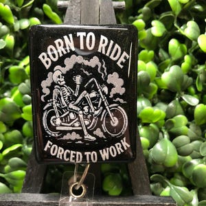May include: Black badge reel with the text "BORN TO RIDE" above a skeleton riding a motorcycle, and "FORCED TO WORK" below. The design is in white. The badge reel has a clear plastic strap and a metal clip. The background is green.