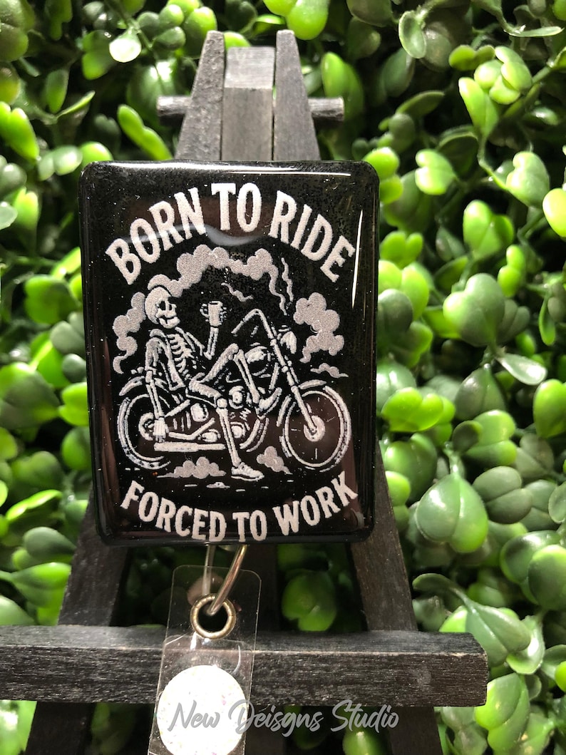 May include: Black badge reel with the text "BORN TO RIDE" above a skeleton riding a motorcycle, and "FORCED TO WORK" below. The design is in white. The badge reel has a clear plastic strap and a metal clip. The background is green.