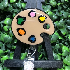 May include: A tan artist's palette badge reel with colorful paint splotches in green, orange, pink, yellow, and blue. The badge reel is on a small black easel with a clear plastic ID holder. The text "New Designs Studio" is visible.