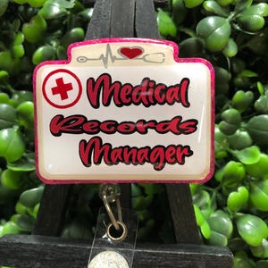 May include: A white and pink Medical Records Manager badge reel with a red cross, heartbeat, and heart design. The badge reel has a glittery pink border and a clear plastic strap with a silver glitter circle.