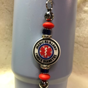 May include: A silver charm with a white, red, and blue medical alert charm that says "MEDICAL ALERT I HAVE DIABETES". The charm is attached to a silver chain with red and blue beads.