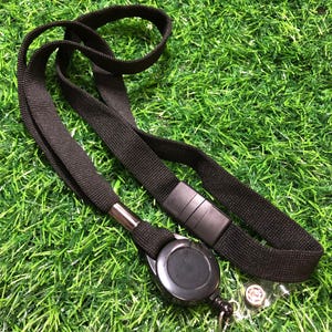May include: Black lanyard with a retractable badge reel and a clear plastic badge holder. The lanyard is made of a woven fabric and has a black plastic clasp. The badge reel is round and black, with a metal clip.