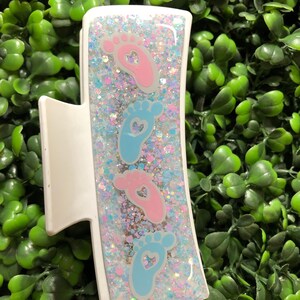 Pu&ograve; includere: Fermaglio bianco per capelli con un design rettangolare riempito di glitter e impronte di bambino. Il fermaglio presenta impronte rosa e blu con ritagli a forma di cuore. Il testo "New Designs Studio" &egrave; in basso.
