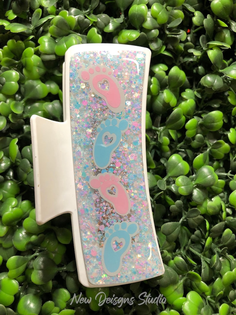 Pu&ograve; includere: Fermaglio bianco per capelli con un design rettangolare riempito di glitter e impronte di bambino. Il fermaglio presenta impronte rosa e blu con ritagli a forma di cuore. Il testo "New Designs Studio" &egrave; in basso.