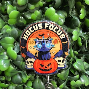 May include: A circular badge reel featuring a black cat wearing an optometry phoropter, sitting on a jack-o'-lantern between two skulls and two crows. The background is an orange sunset with bats and leaves, and the text HOCUS FOCUS is printed.