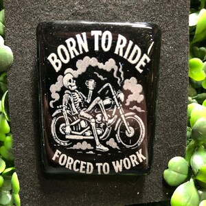 May include: A black rectangular resin piece with the words "BORN TO RIDE" and "FORCED TO WORK" in white. A skeleton is riding a motorcycle, holding a mug. The background is black with a glittery effect.