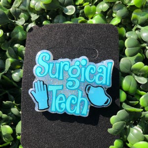 May include: A light blue and white glittery "Surgical Tech" badge with a glove and mask icon. The badge is on a black background and surrounded by green leaves.