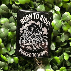 May include: Black square badge reel with the text "BORN TO RIDE FORCED TO WORK" in white. The design features a skeleton riding a motorcycle, holding a cup. The badge reel has a clear plastic clip and a retractable cord.