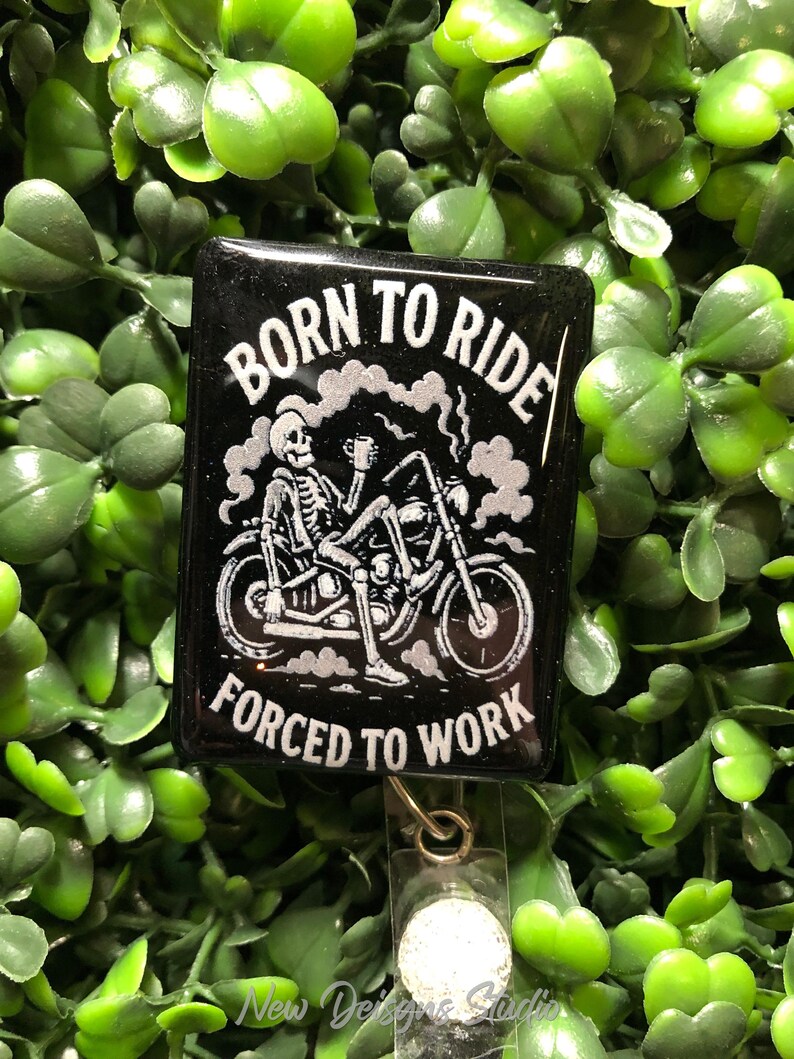 May include: Black square badge reel with the text "BORN TO RIDE FORCED TO WORK" in white. The design features a skeleton riding a motorcycle, holding a cup. The badge reel has a clear plastic clip and a retractable cord.