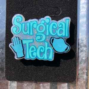 May include: A badge with the words "Surgical Tech" in light blue, along with a blue surgical glove and mask. The badge is on a black background and has a silver clip at the top.