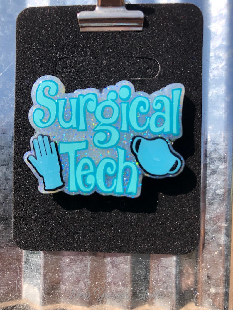 May include: A badge with the words "Surgical Tech" in light blue, along with a blue surgical glove and mask. The badge is on a black background and has a silver clip at the top.