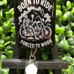 May include: Black badge reel with the text "BORN TO RIDE FORCED TO WORK" and a skeleton riding a motorcycle. The design is in white, with smoke and a coffee cup. The reel has a clear plastic strap and a metal clip.