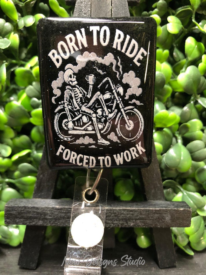 May include: Black badge reel with the text "BORN TO RIDE FORCED TO WORK" and a skeleton riding a motorcycle. The design is in white, with smoke and a coffee cup. The reel has a clear plastic strap and a metal clip.