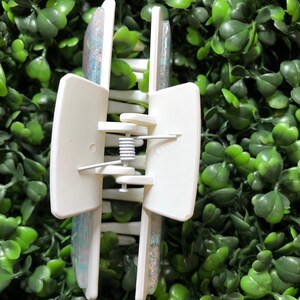 Pu&ograve; includere: Un fermaglio per capelli rettangolare bianco con un design glitterato e iridescente. Il fermaglio ha un meccanismo a molla ed &egrave; posto su uno sfondo di fogliame verde. Il testo "New Designs Studio" &egrave; visibile.