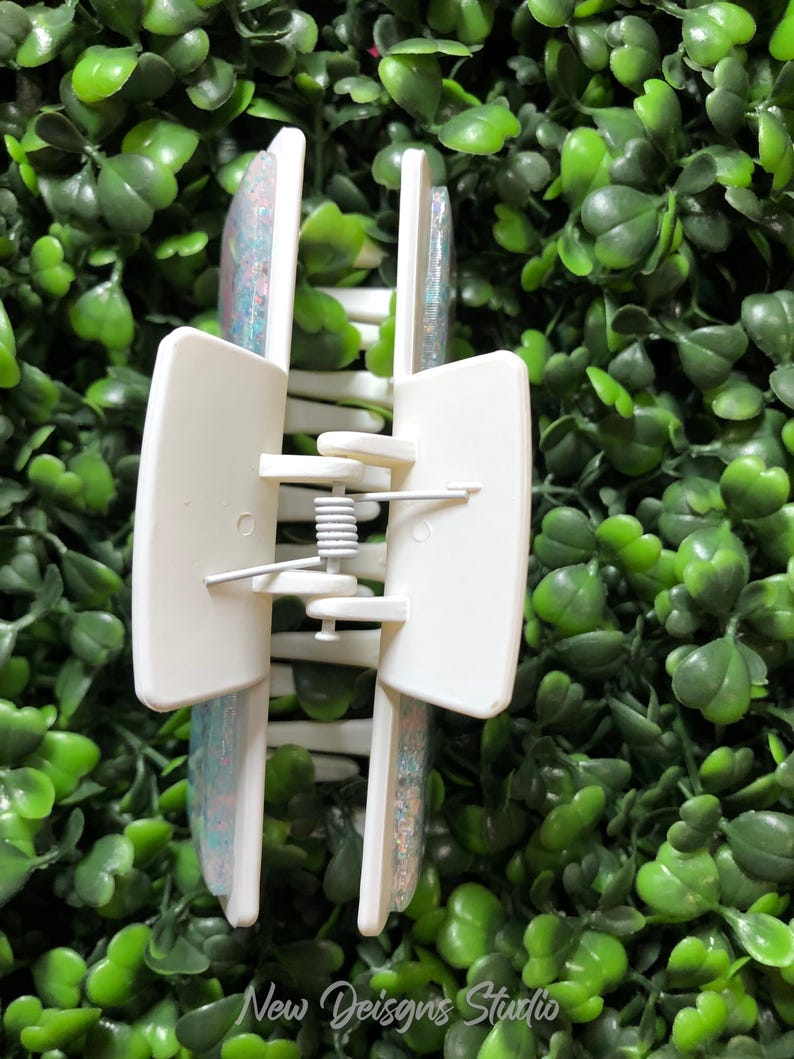 Pu&ograve; includere: Un fermaglio per capelli rettangolare bianco con un design glitterato e iridescente. Il fermaglio ha un meccanismo a molla ed &egrave; posto su uno sfondo di fogliame verde. Il testo "New Designs Studio" &egrave; visibile.