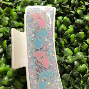 Pu&ograve; includere: Fermaglio bianco per capelli con un design rettangolare con impronte di piedi di bambino rosa e blu con ritagli a forma di cuore. Il fermaglio &egrave; riempito di glitter e il testo "New Designs Studio" &egrave; visibile.
