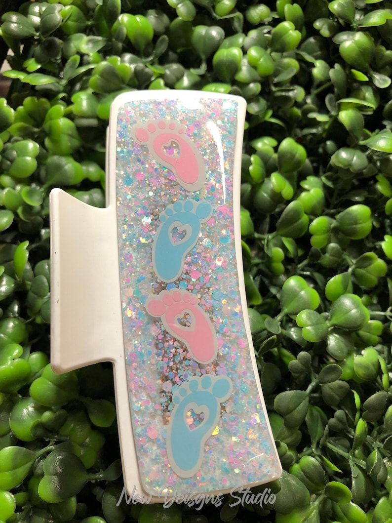 Pu&ograve; includere: Fermaglio bianco per capelli con un design rettangolare con impronte di piedi di bambino rosa e blu con ritagli a forma di cuore. Il fermaglio &egrave; riempito di glitter e il testo "New Designs Studio" &egrave; visibile.