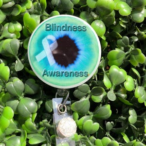 May include: A round badge with the words "Blindness Awareness" and a light blue ribbon design over a blue and black eye graphic. The badge is attached to a clear plastic retractable reel with a decorative, sparkly button.