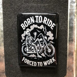 May include: Black rectangular magnet with the text "BORN TO RIDE FORCED TO WORK" in white. The graphic shows a skeleton riding a motorcycle, holding a cup. The magnet is on a black felt-like surface.