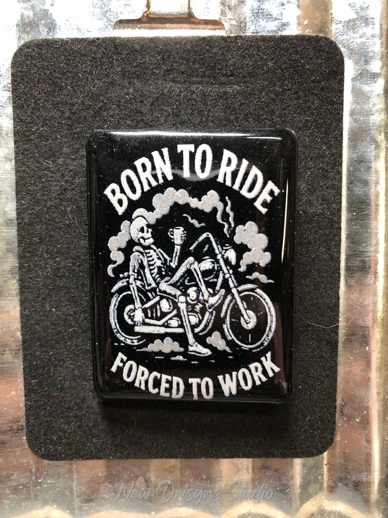 May include: Black rectangular magnet with the text "BORN TO RIDE FORCED TO WORK" in white. The graphic shows a skeleton riding a motorcycle, holding a cup. The magnet is on a black felt-like surface.