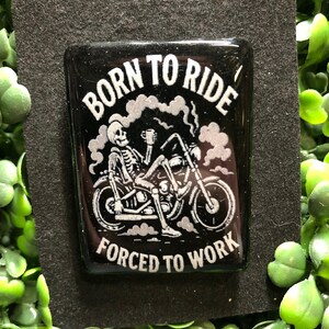 May include: A black rectangular pin with the words "BORN TO RIDE" above and "FORCED TO WORK" below a skeleton riding a motorcycle. The skeleton is holding a cup, and smoke is rising from the motorcycle.