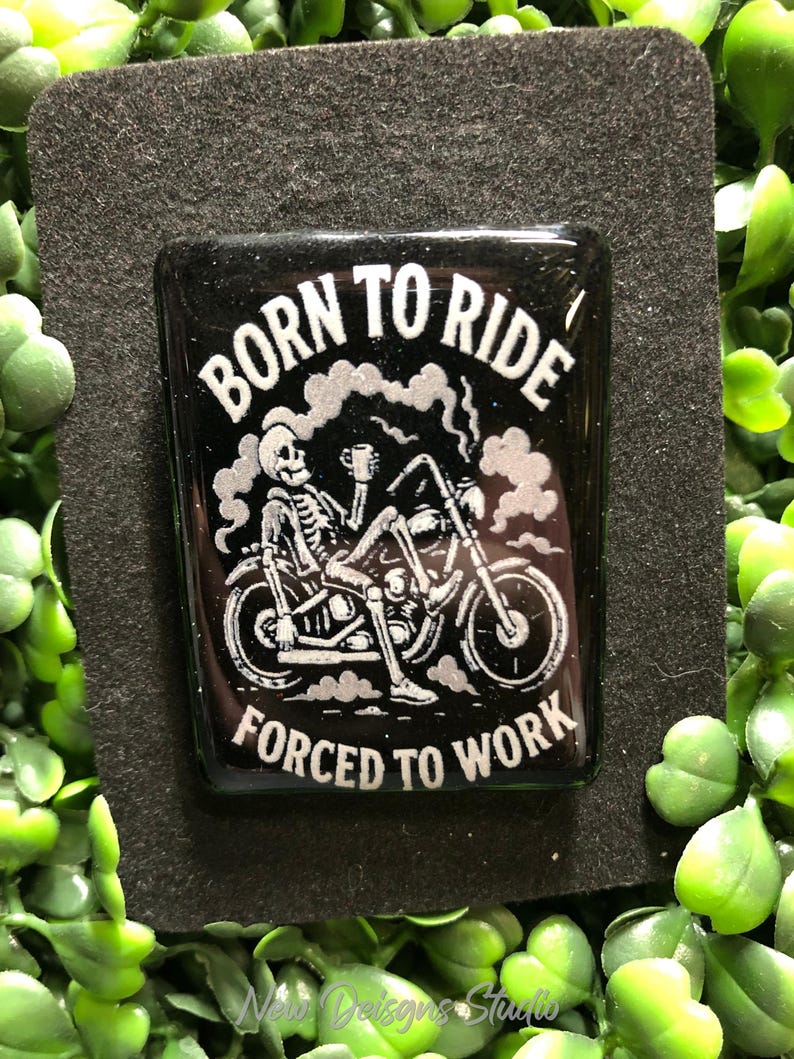 May include: A black rectangular pin with the words "BORN TO RIDE" above and "FORCED TO WORK" below a skeleton riding a motorcycle. The skeleton is holding a cup, and smoke is rising from the motorcycle.