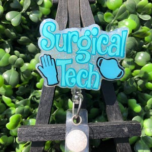May include: A badge reel with the words "Surgical Tech" in turquoise lettering, a blue glove, and a surgical mask. The badge is on a black easel with a clear plastic holder and a white button.