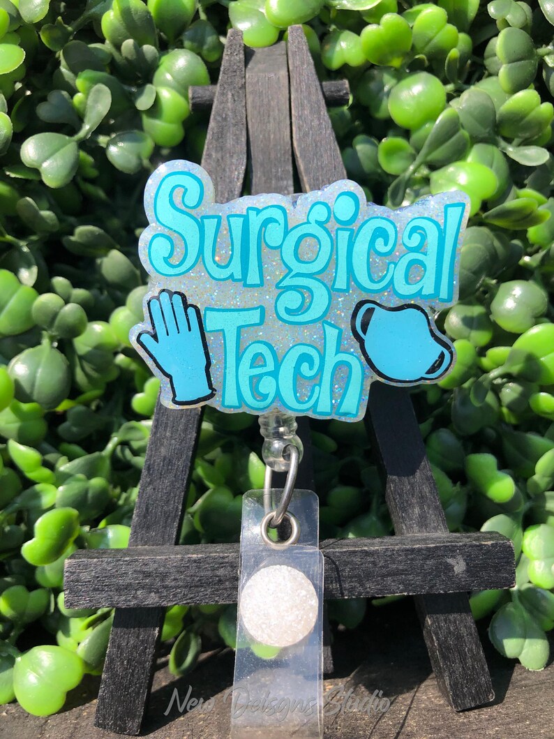 May include: A badge reel with the words "Surgical Tech" in turquoise lettering, a blue glove, and a surgical mask. The badge is on a black easel with a clear plastic holder and a white button.