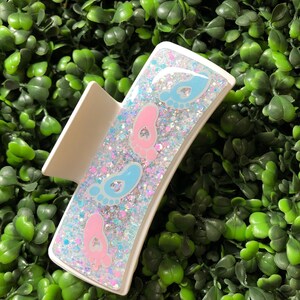 Pu&ograve; includere: Un fermaglio per capelli bianco con un design rettangolare riempito di glitter e impronte di piedi di bambino. Il fermaglio presenta impronte rosa e blu, con il testo "New Designs Studio" in basso.
