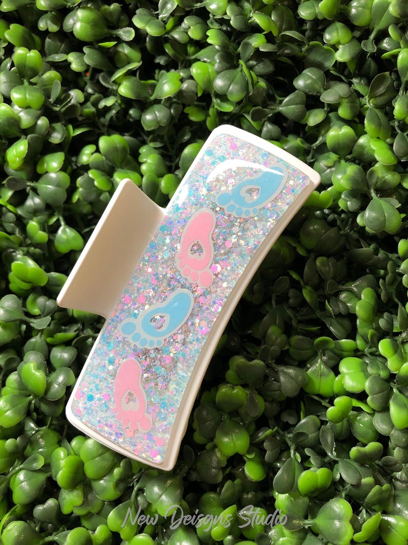 Pu&ograve; includere: Un fermaglio per capelli bianco con un design rettangolare riempito di glitter e impronte di piedi di bambino. Il fermaglio presenta impronte rosa e blu, con il testo "New Designs Studio" in basso.