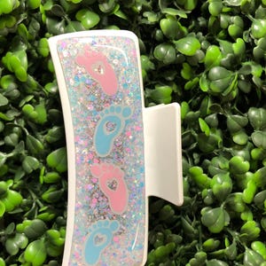 Pu&ograve; includere: Fermaglio bianco con un design rettangolare con impronte di piedi di bambino rosa e blu con ritagli a forma di cuore, su uno sfondo glitterato. La clip ha il testo "New Designs Studio" in basso.
