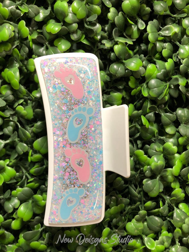 Pu&ograve; includere: Fermaglio bianco con un design rettangolare con impronte di piedi di bambino rosa e blu con ritagli a forma di cuore, su uno sfondo glitterato. La clip ha il testo "New Designs Studio" in basso.
