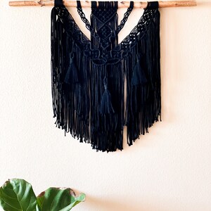 May include: Black macrame wall hanging with a geometric design and fringe. The hanging is attached to a natural wood dowel.
