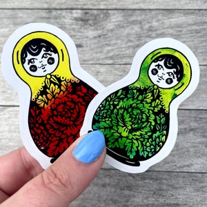 May include: Two nesting doll stickers with a white background. The doll on the left has a yellow head and red, orange, and yellow floral pattern. The doll on the right has a green head and green floral pattern.